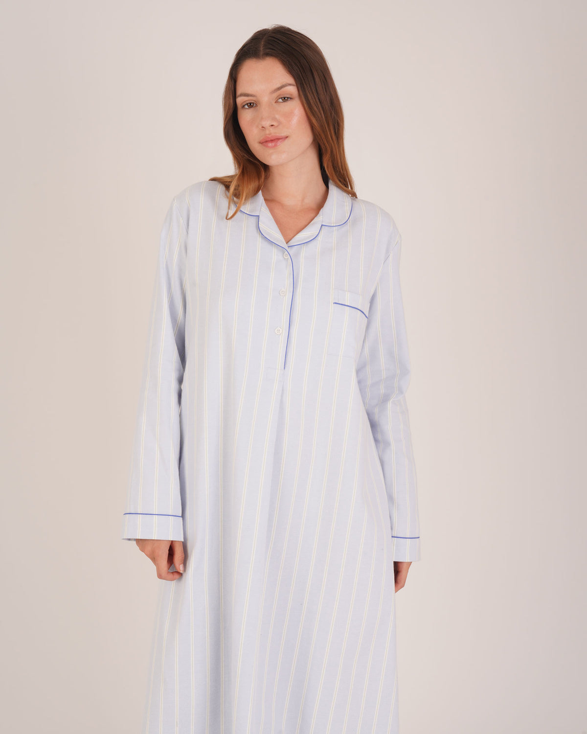 Bonsoir of London Women’s Isla Blue White Stripe Brushed Cotton Nightshirt, featuring a blue and white striped design, button placket, shirt-tail hem, piping detail, cuffs and breast pocket, half body shot on a model.