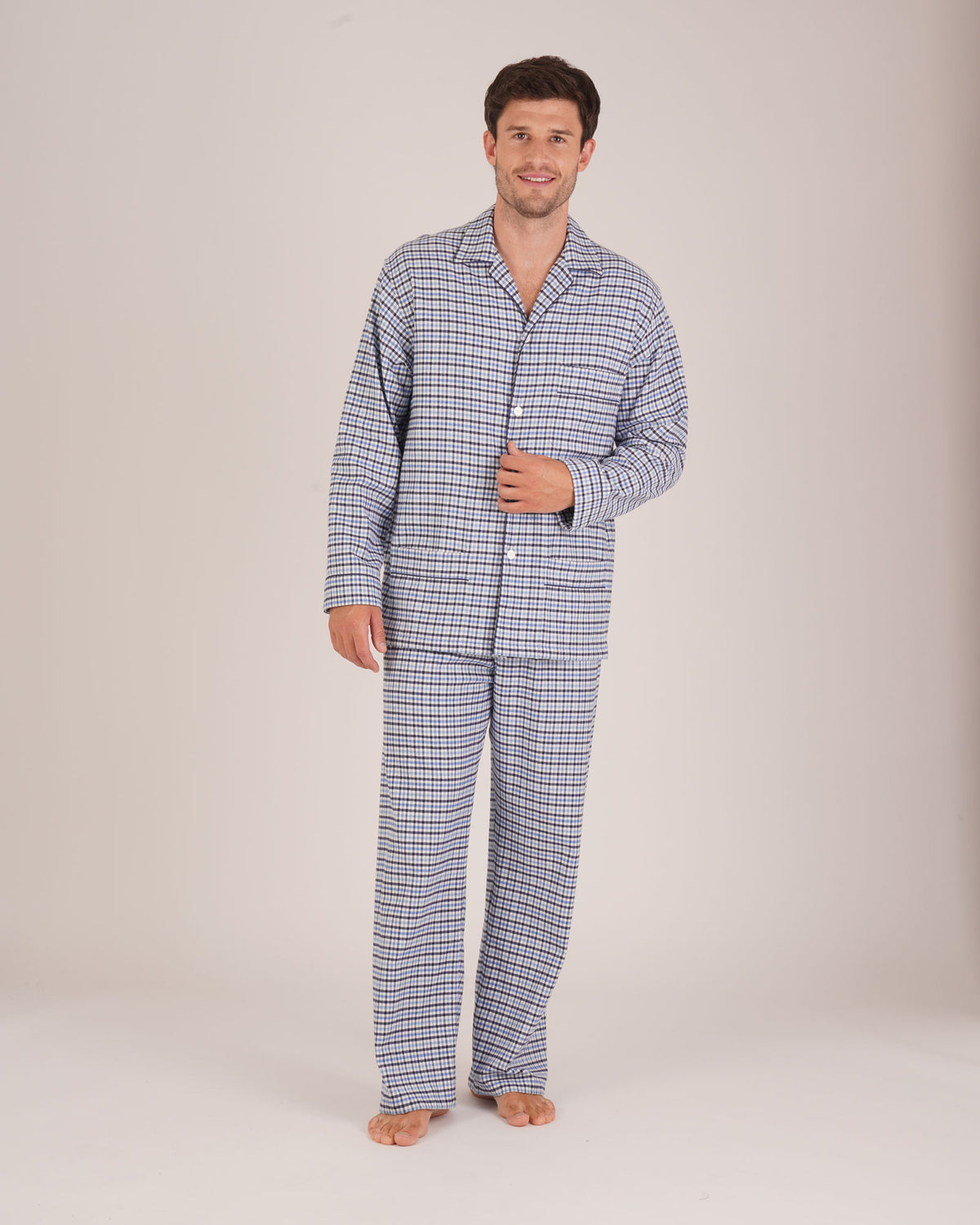 Bonsoir of London Men’s Arthur Blue Check Brushed Cotton Pyjamas, featuring a blue, pale blue, white and navy check design, three jacket pockets, elasticated-waist trousers, button fastening and concealed button fly, full body shot on a model.