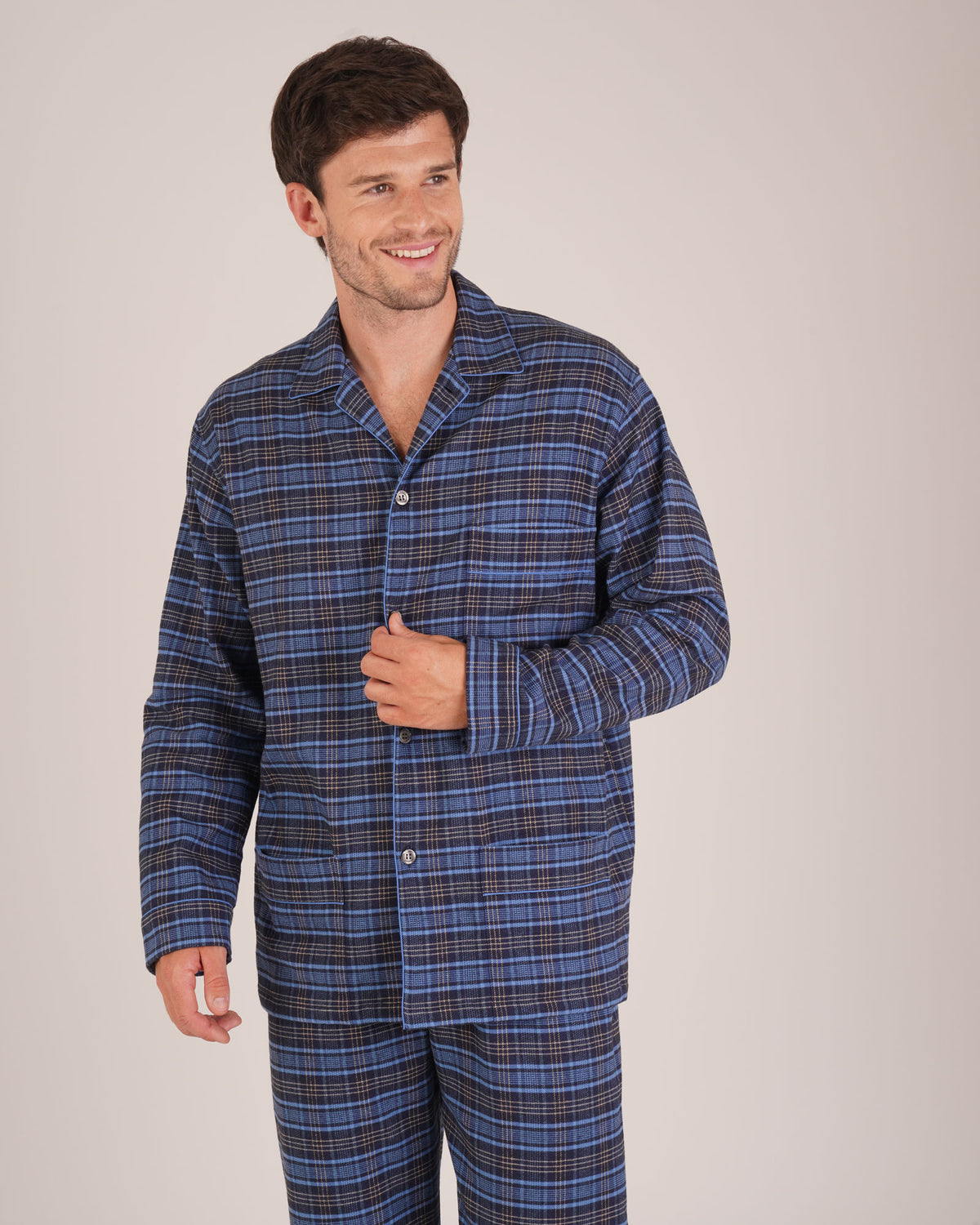 Bonsoir of London Men’s Theodore Blue Plaid Brushed Cotton Pyjamas, featuring a blue and navy plaid design with subtle gold detailing, three jacket pockets, elasticated-waist trousers, button fastening and concealed button fly, half body shot on a model.