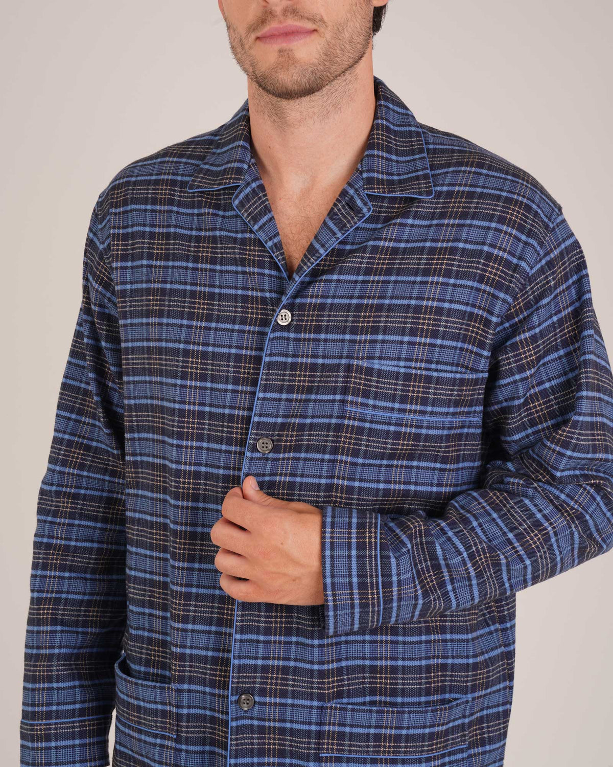 Bonsoir of London Men’s Theodore Blue Plaid Brushed Cotton Pyjamas, featuring a blue and navy plaid design with subtle gold detailing, three jacket pockets, elasticated-waist trousers, button fastening and concealed button fly, close up shot on a model.