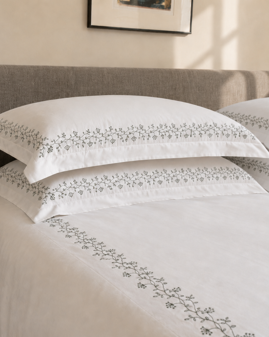 Bonsoir of London white cotton embroidered bedding with a green floral design, pictured on a bed with a grey fabric headboard.