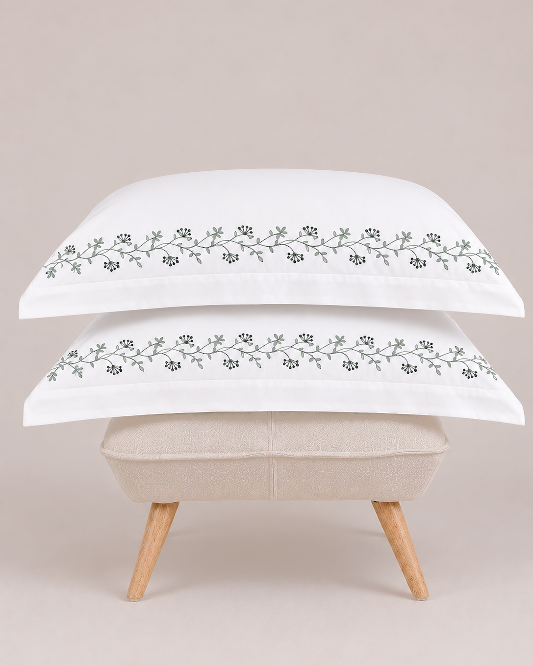 Bonsoir of London two white pillows with green floral embroidery on a beige ottoman against a light studio background.