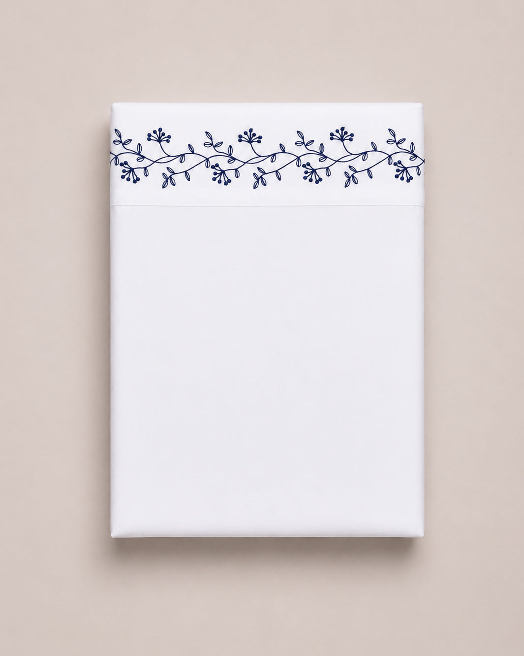 Bonsoir of London cotton white flat sheet with navy floral embroidery, folded on a beige studio background.