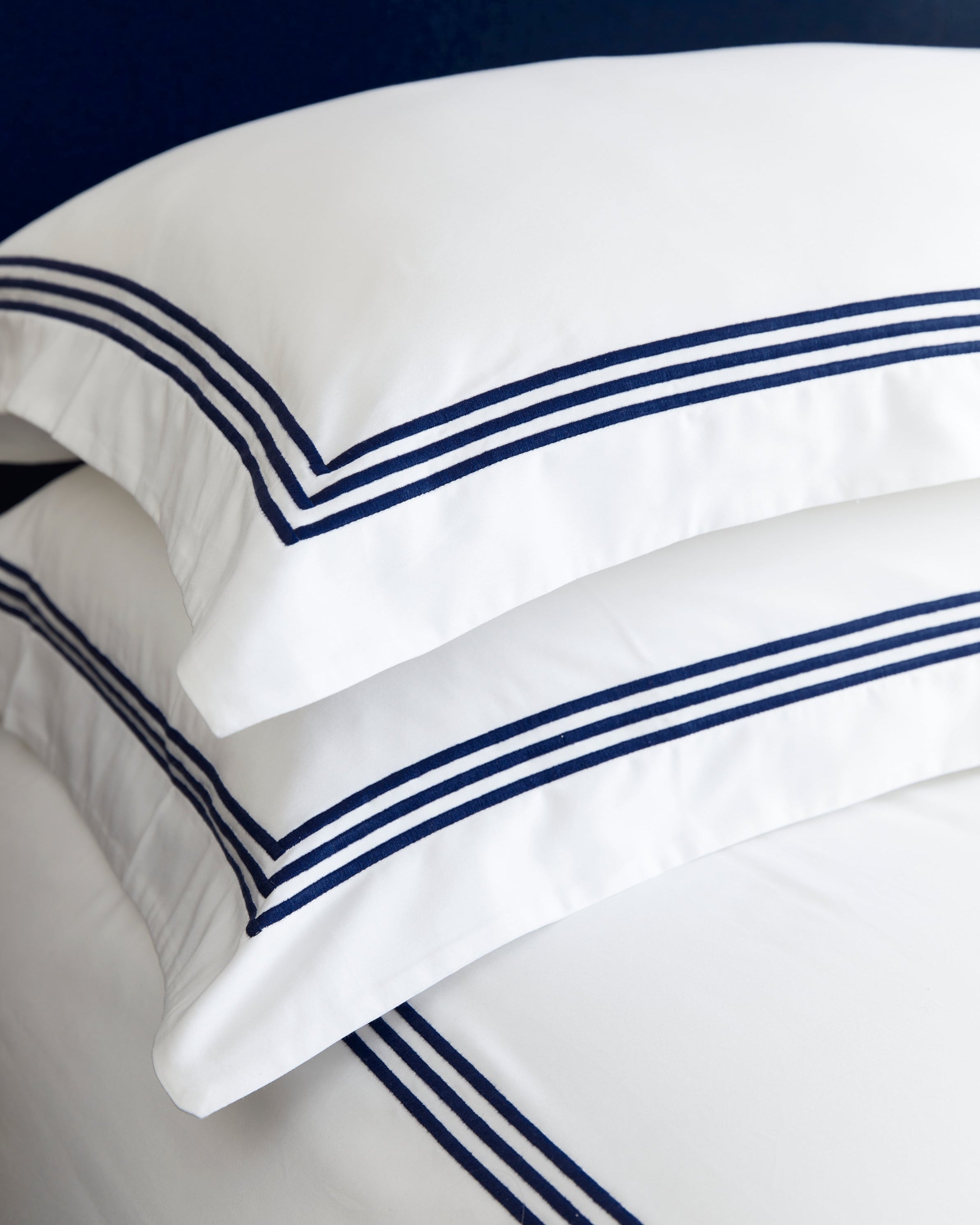 Bonsoir of London white cotton sateen pillowcases with navy blue triple-stripe embroidery, placed on a bed with a navy headboard, close-up shot.