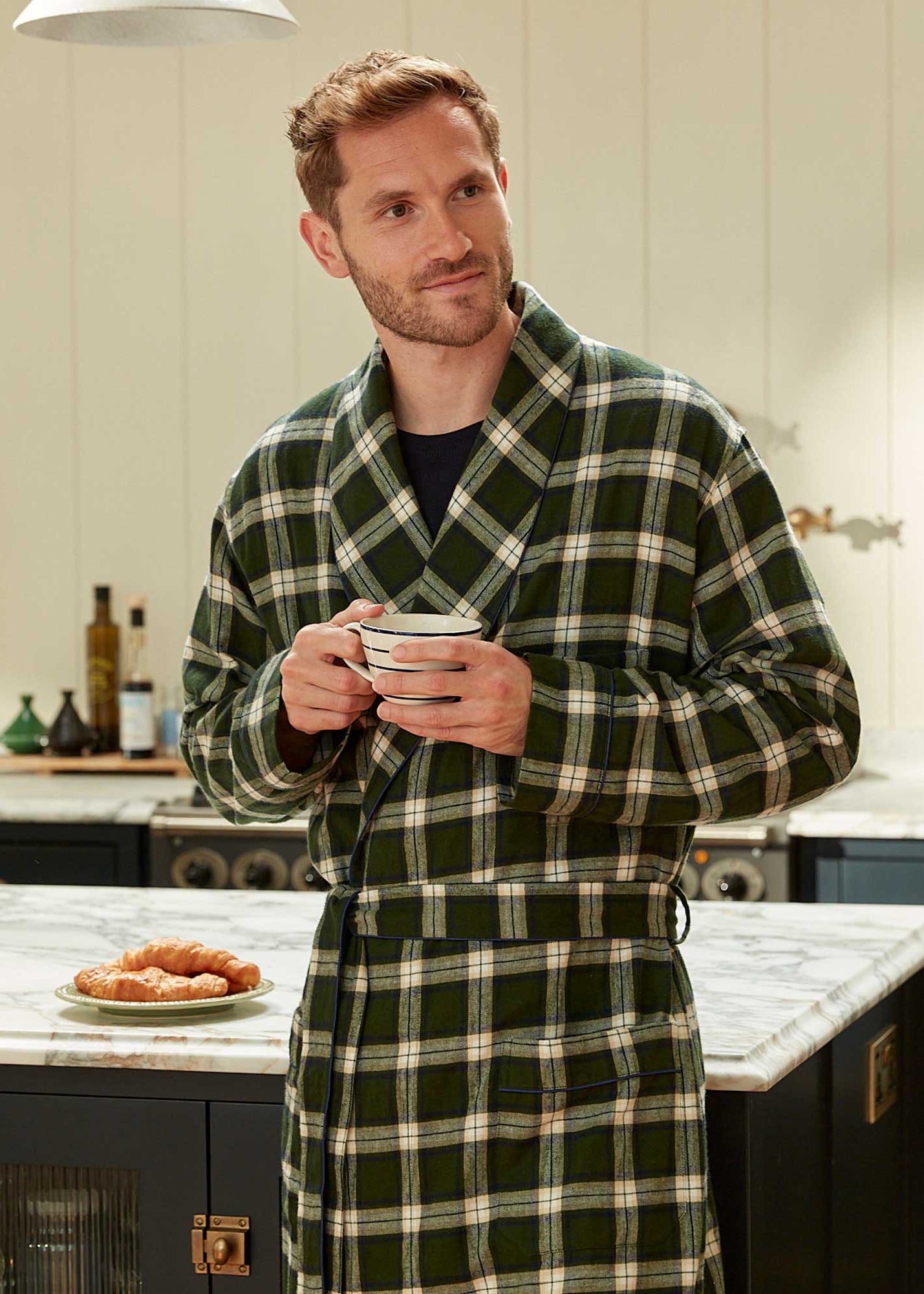 Man wearing a green plaid robe holding a mug in a kitchen.
