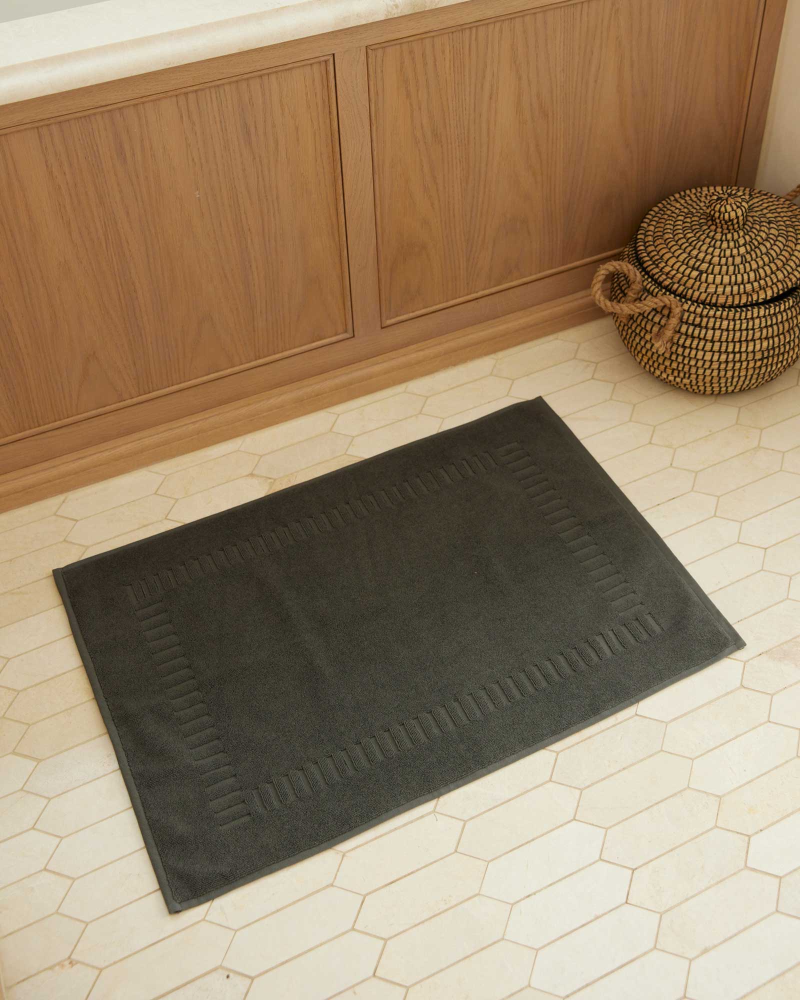 Bonsoir of London 100% cotton bath mat in charcoal grey, full lifestyle shot on a tile floor.
