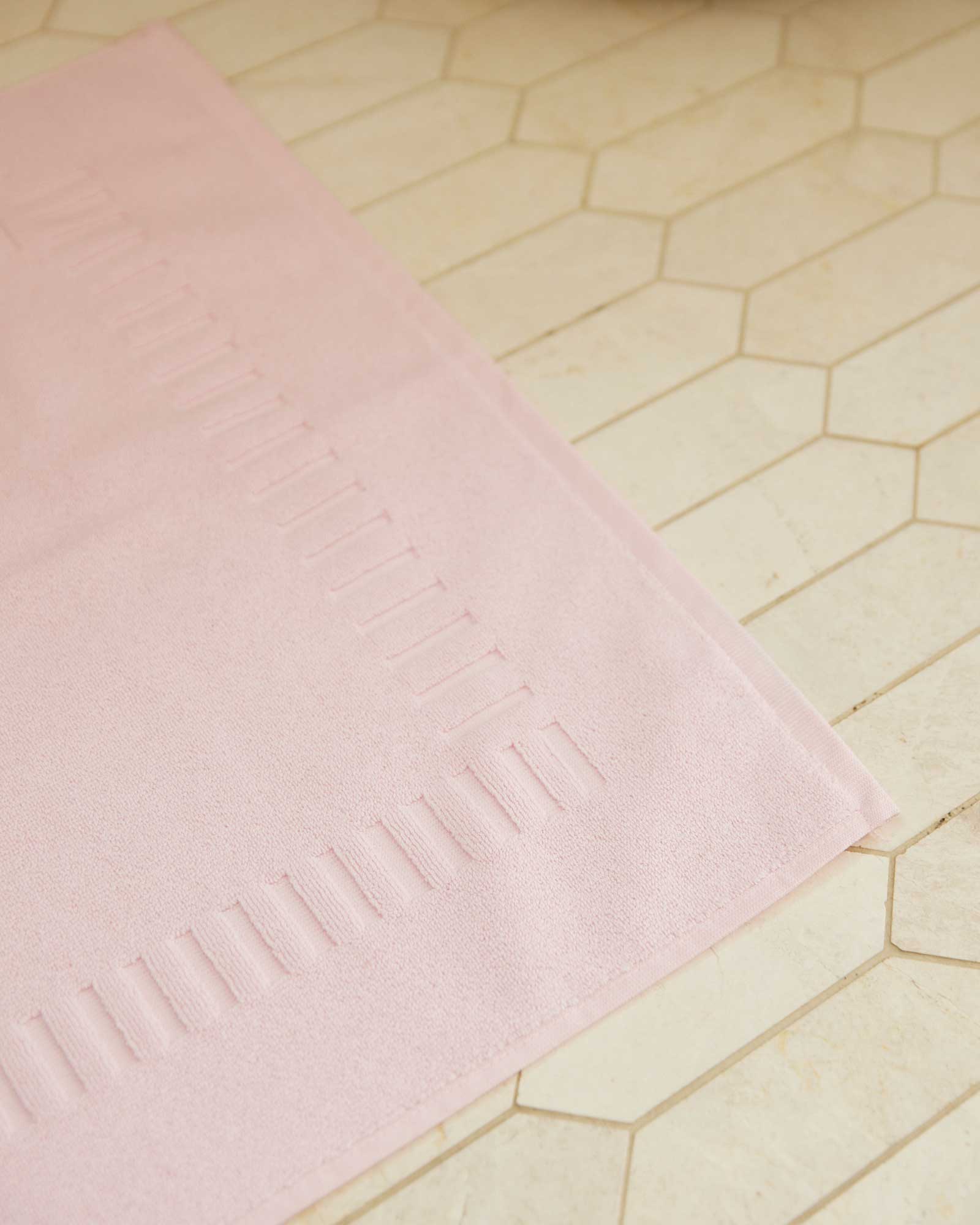 Bonsoir of London 100% Cotton Bathmat in pink, detail close-up on a beige tile floor.