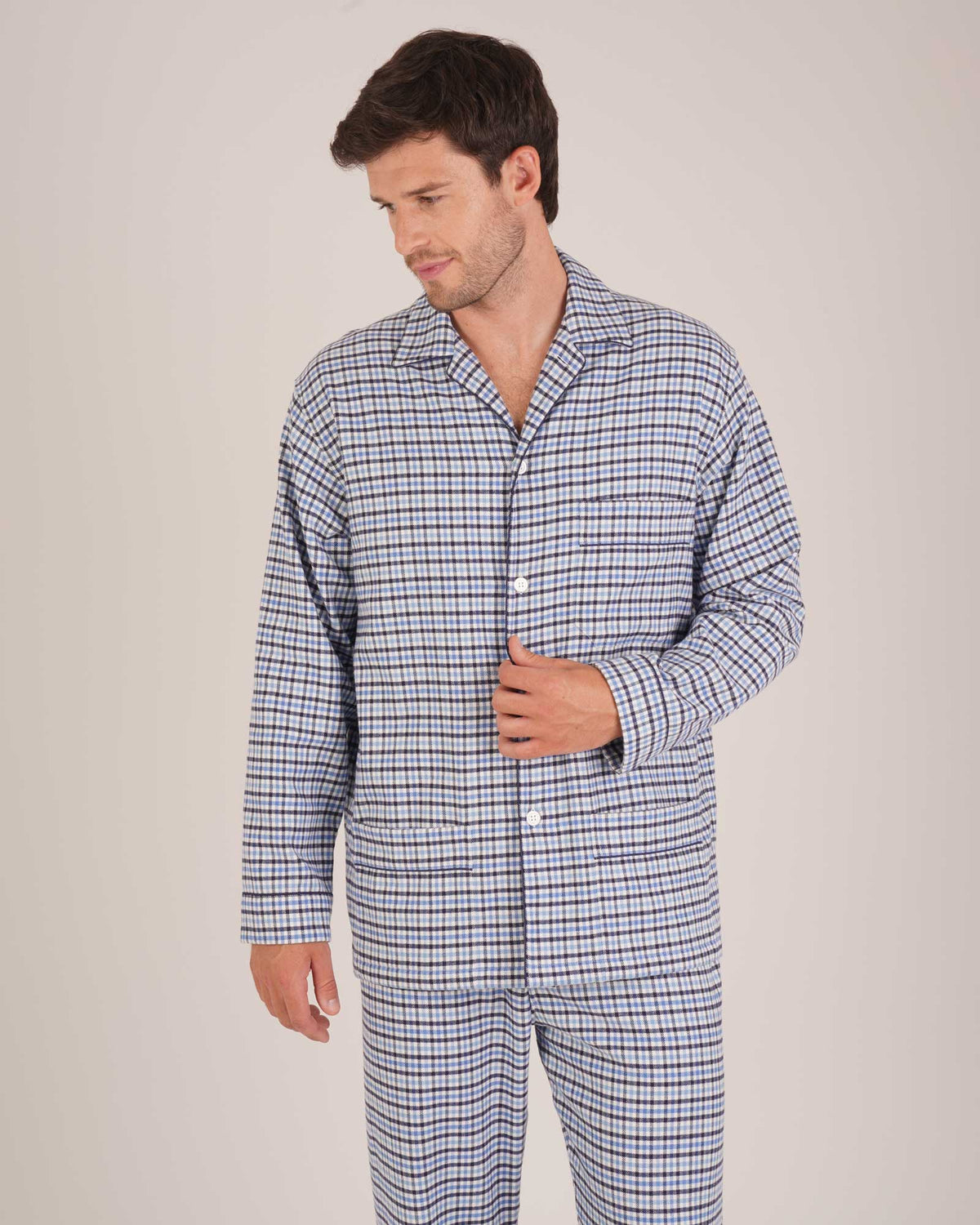 Bonsoir of London Men’s Arthur Blue Check Brushed Cotton Pyjamas, featuring a blue, pale blue, white and navy check design, three jacket pockets, elasticated-waist trousers, button fastening and concealed button fly, half body shot on a model.