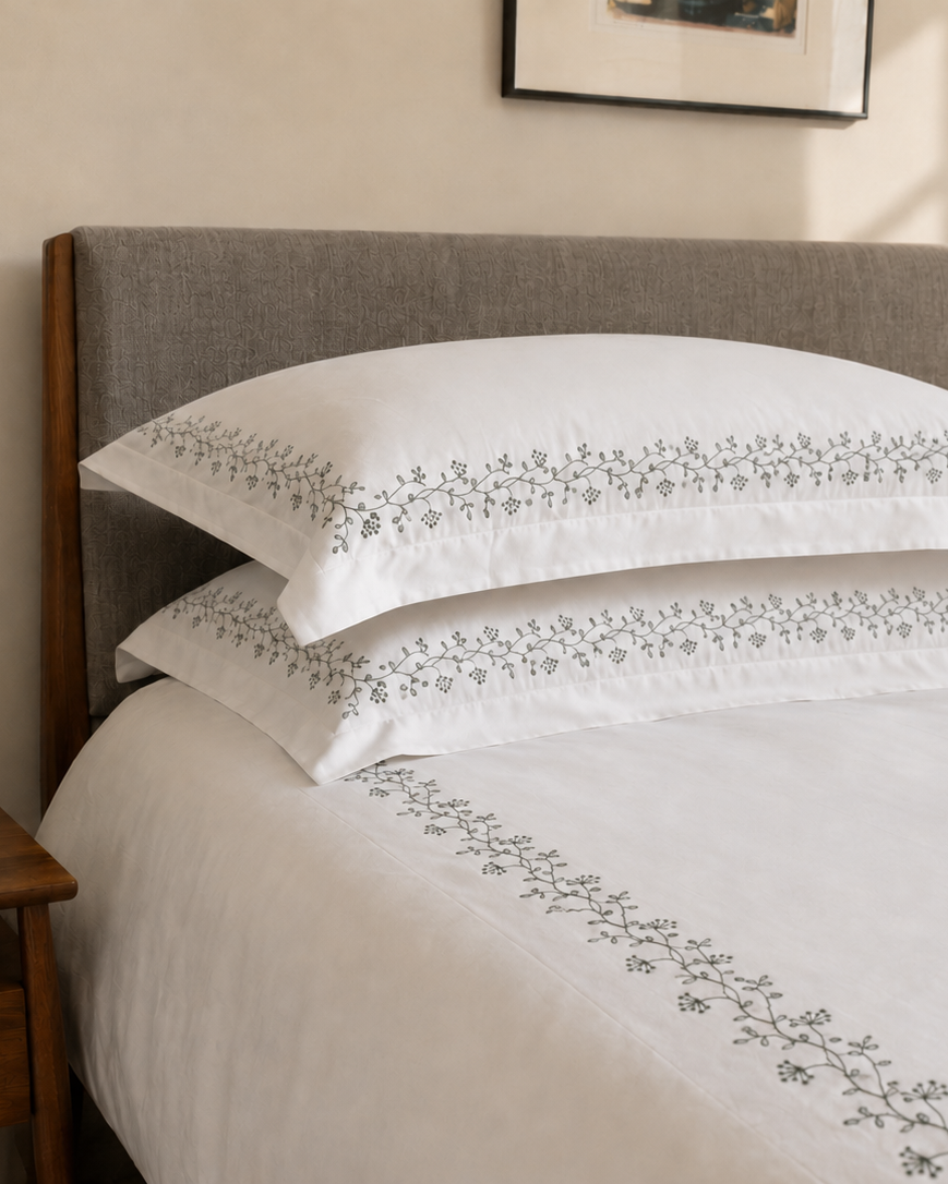 Bonsoir of London white bedding with green floral embroidery, on a bed with a wooden nightstand and decorative items.