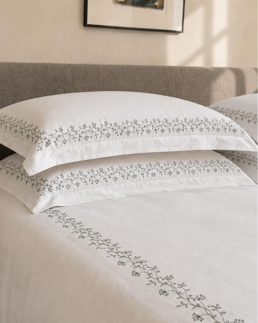 Bonsoir of London white cotton embroidered bedding with a green floral design, pictured on a bed with a grey fabric headboard.
