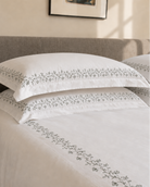 Bonsoir of London white cotton embroidered bedding with a green floral design, pictured on a bed with a grey fabric headboard.