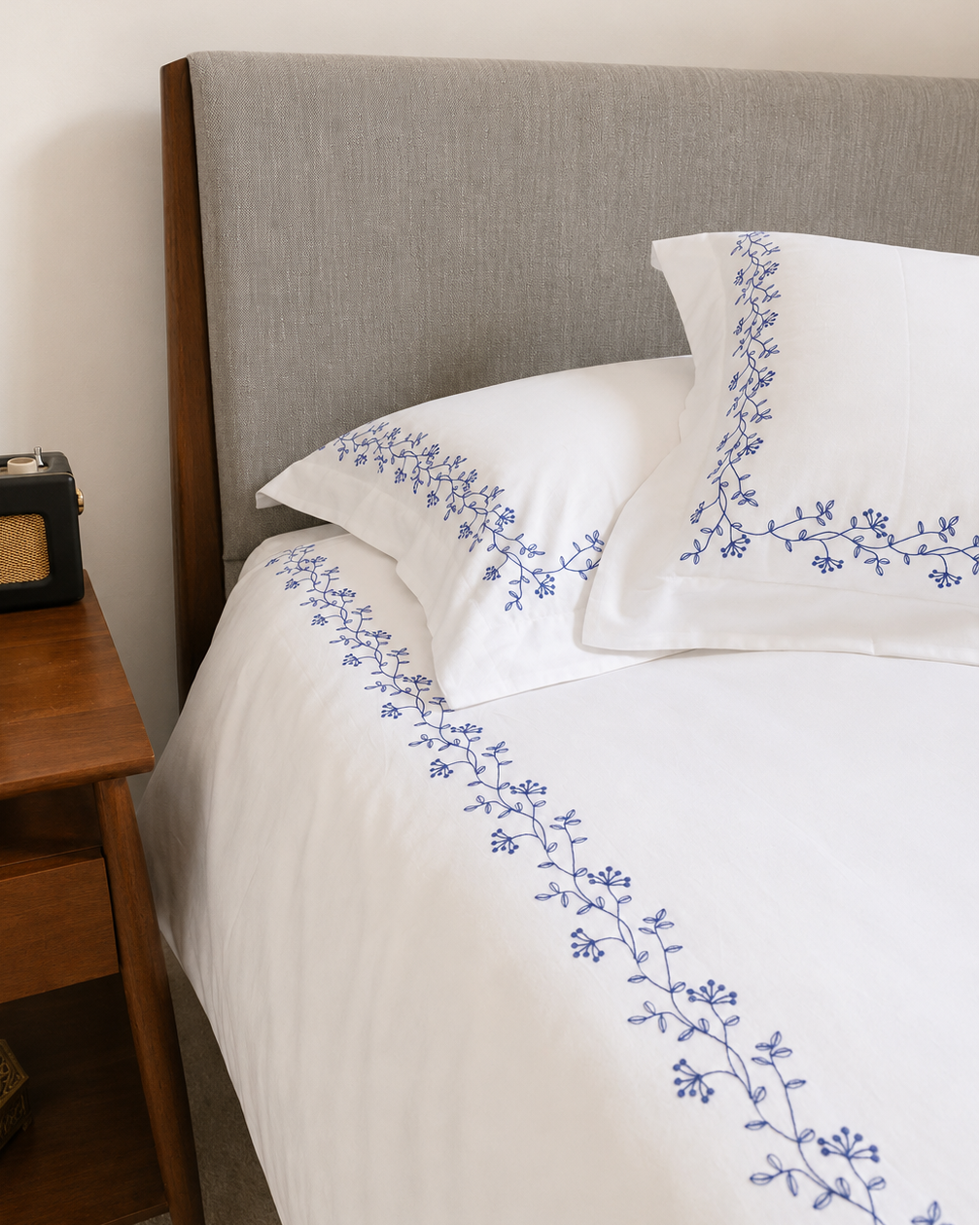 Bonsoir of London Kensington bedding with navy embroidery shot on a bed with a gray headboard, next to a wooden nightstand.