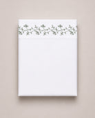 Bonsoir of London cotton white flat sheet with green floral embroidery, folded on a beige studio background.