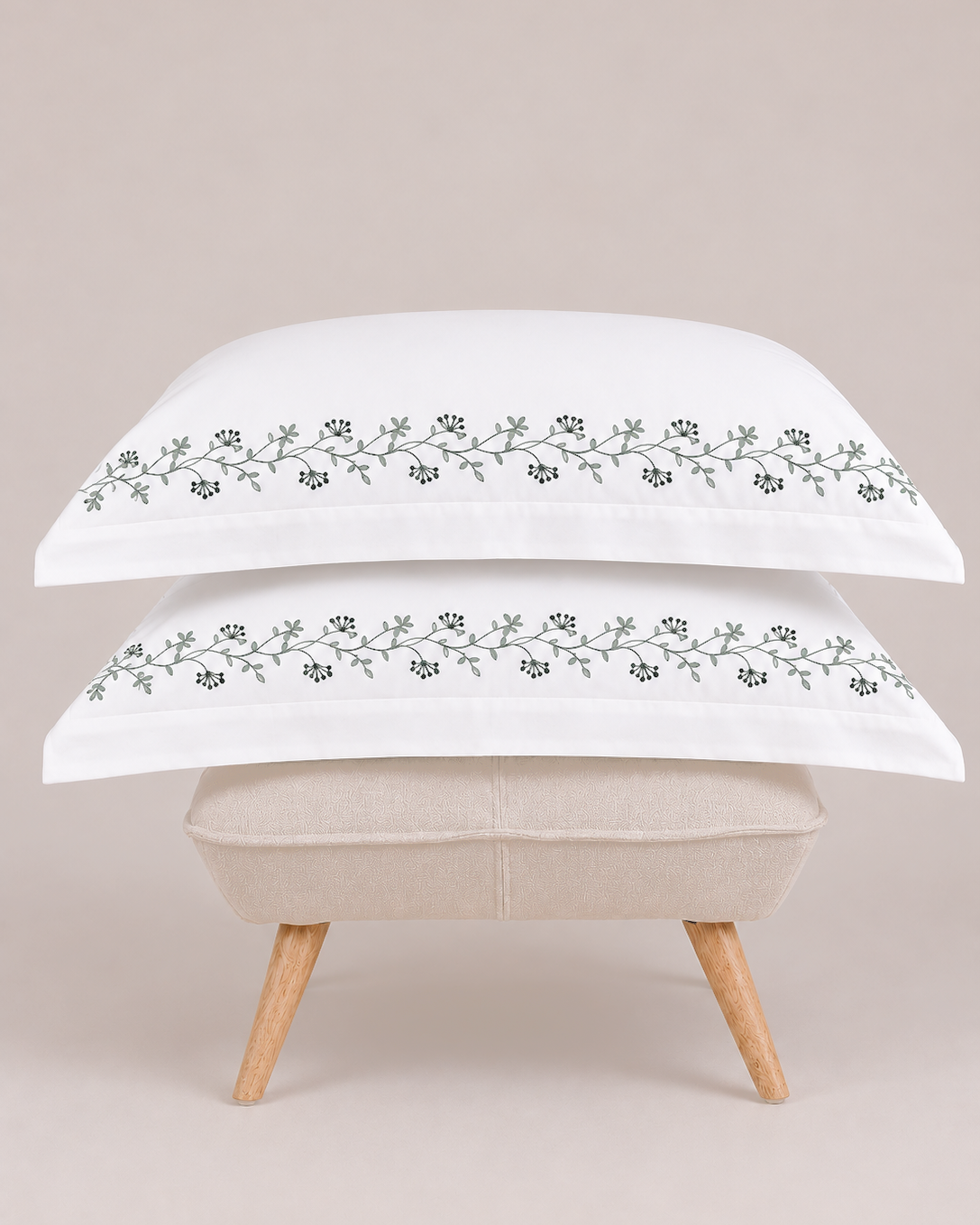 Bonsoir of London two white pillows with green floral embroidery on a beige ottoman against a light studio background.