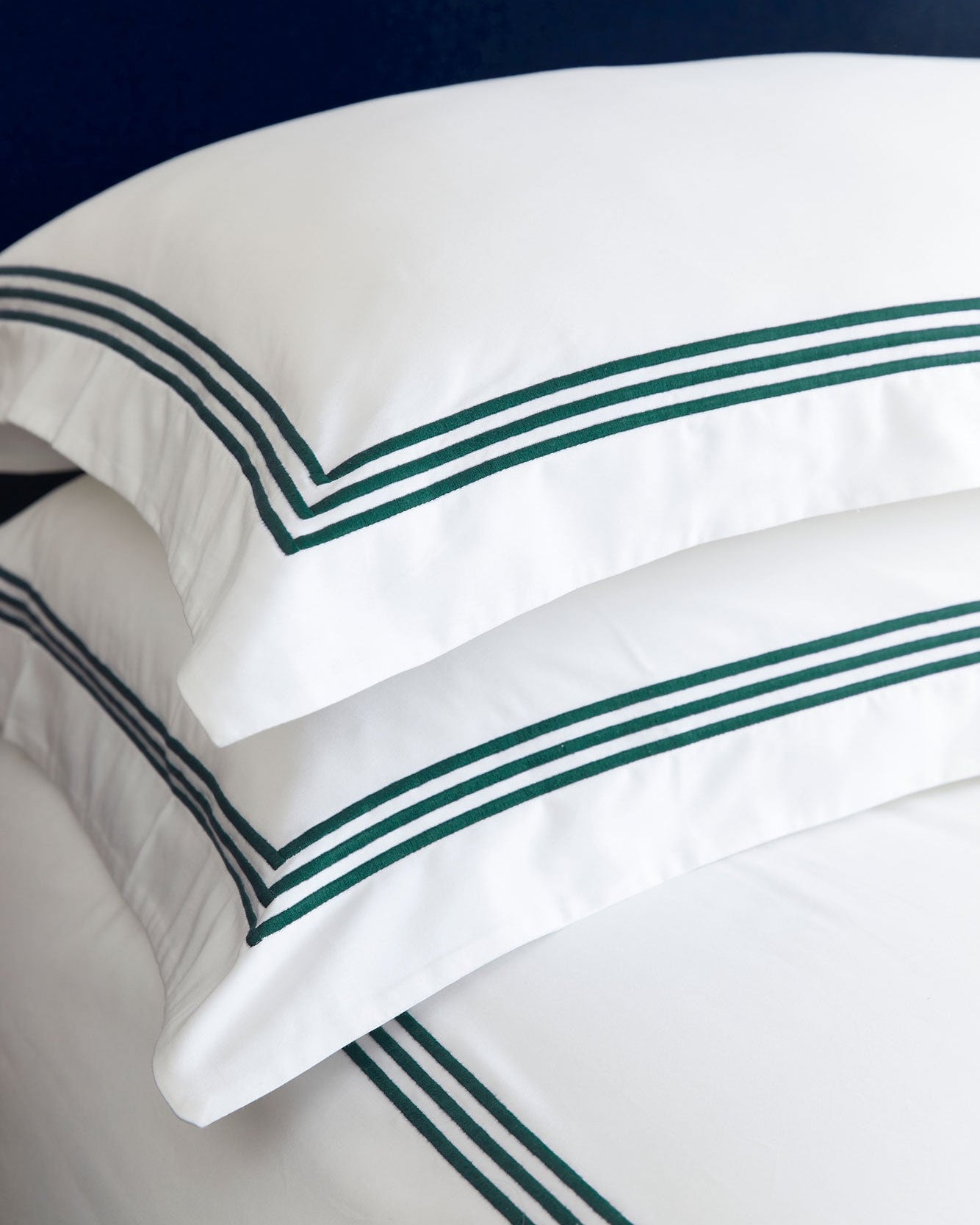 Bonsoir of London white cotton pillows with three green stripes of embroidery, close-up on a bed against a blue background.