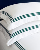 Bonsoir of London white cotton pillows with three green stripes of embroidery, close-up on a bed against a blue background.