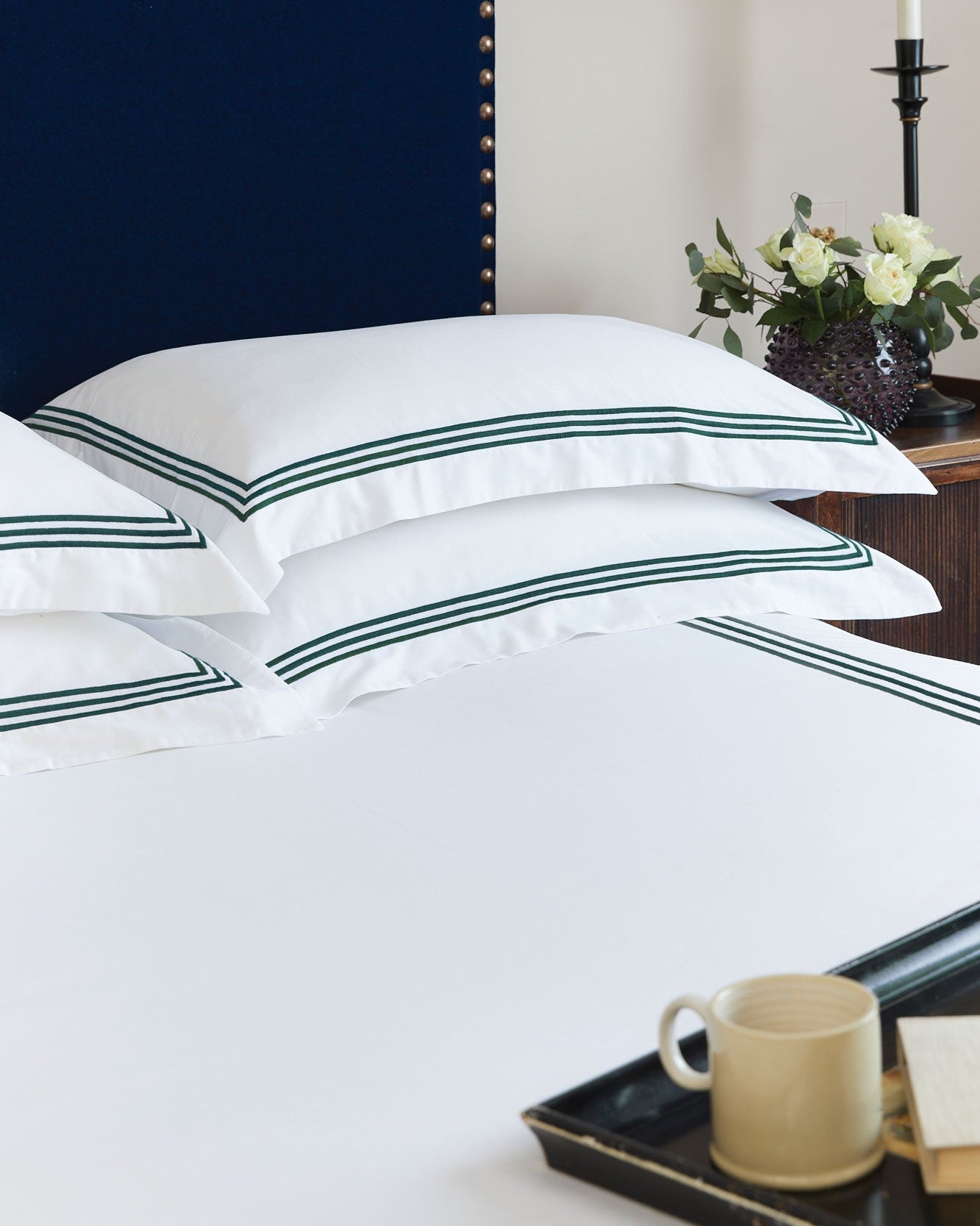 Bonsoir of London white cotton sateen bed sheets with green stripes on a bed, with a nightstand and lamp in the background.