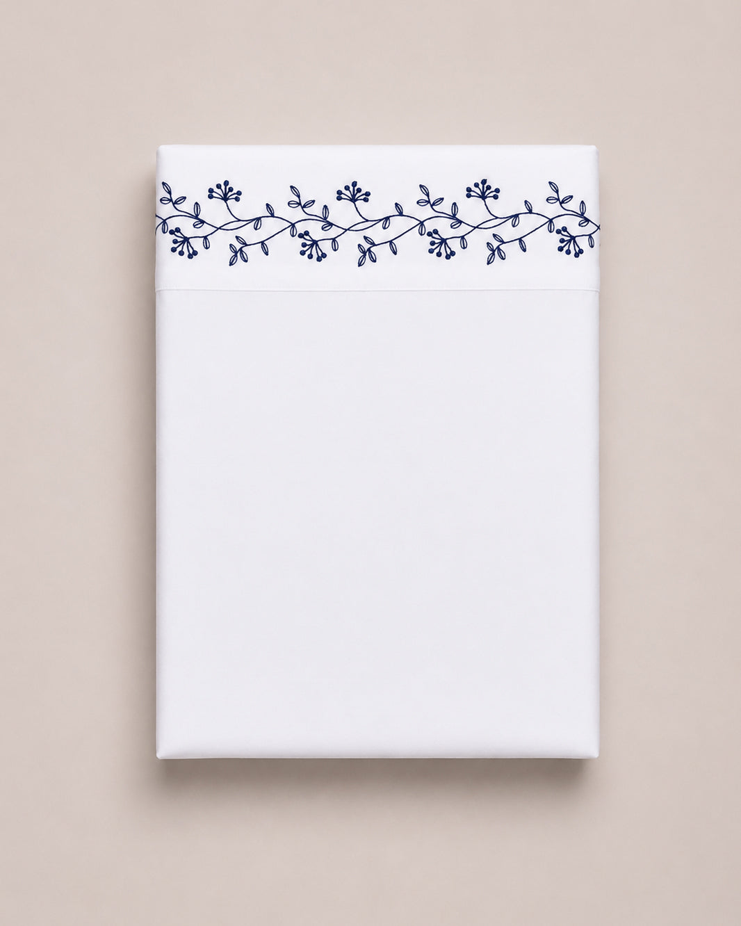 Bonsoir of London cotton white flat sheet with navy floral embroidery, folded on a beige studio background.