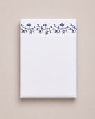 Bonsoir of London cotton white flat sheet with navy floral embroidery, folded on a beige studio background.